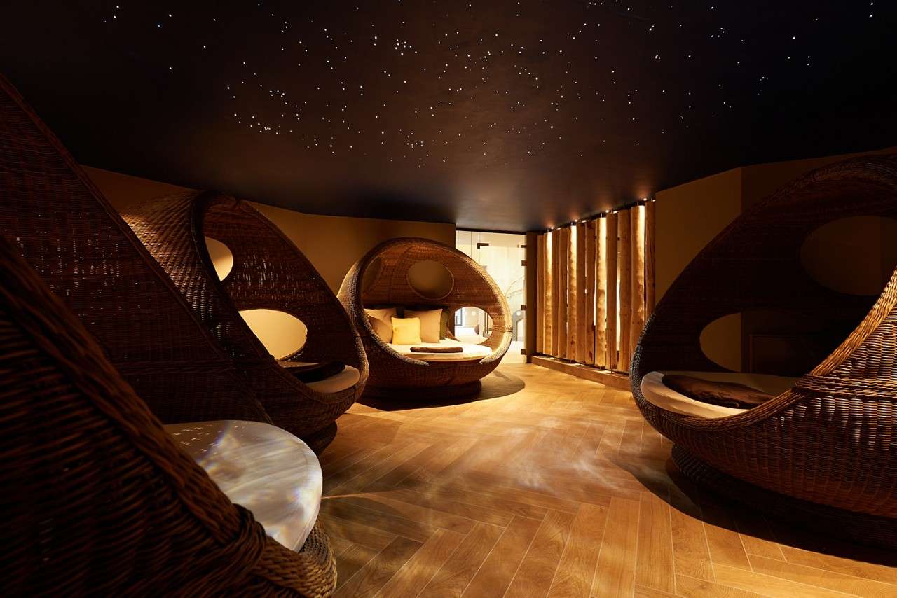 Egg-shaped wicker lounge pods cradle cushions, inviting rest, within a dim spa lounge; warm wood floors, a slatted timber wall, and a starry fiber‑optic ceiling surround a glass-doored entrance.
