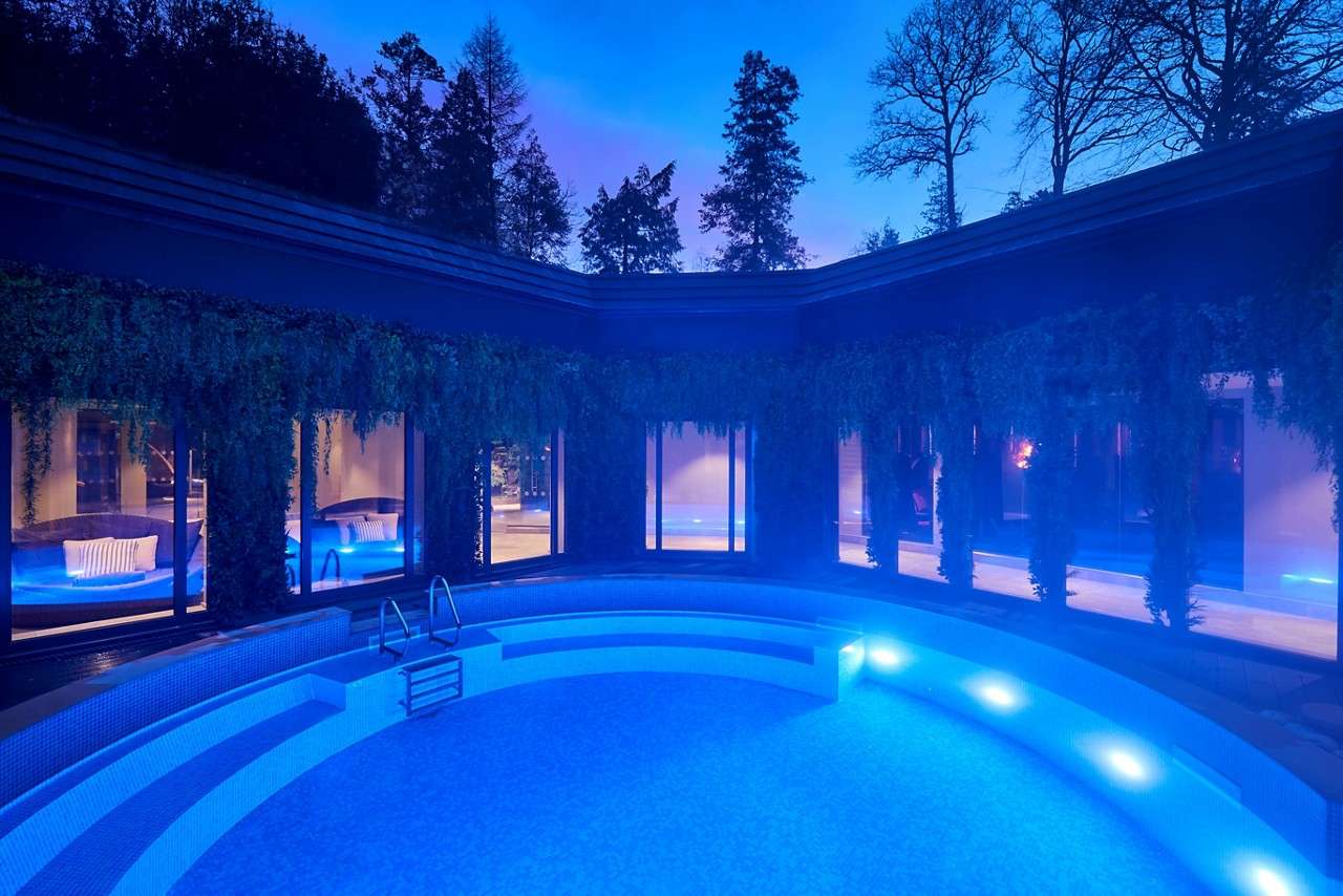 Curved swimming pool glows blue, lights washing steps and benches. Surrounding building with large windows and hanging greenery encloses a courtyard, while tall trees silhouette against twilight sky.