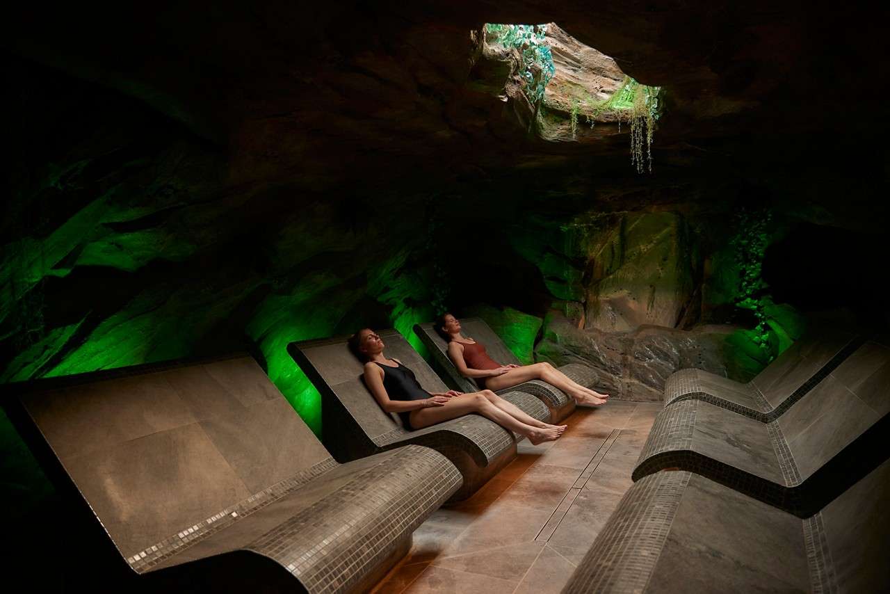 Two spa-goers recline motionless on curved, tiled loungers, relaxing. Around them, a cave-like room with rock walls glows green from uplights, while a small skylight overhead reveals plants and daylight.
