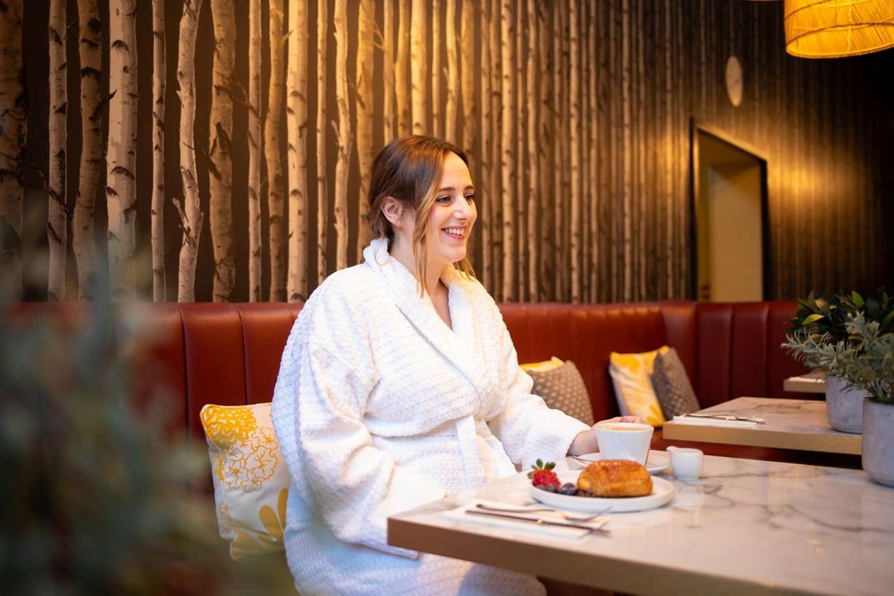 Woman in a white bathrobe smiles while eating breakfast—croissant, berries, and coffee—at a marble table. She sits in a red booth with pillows, birch-tree wallpaper, warm lighting, and potted plants.