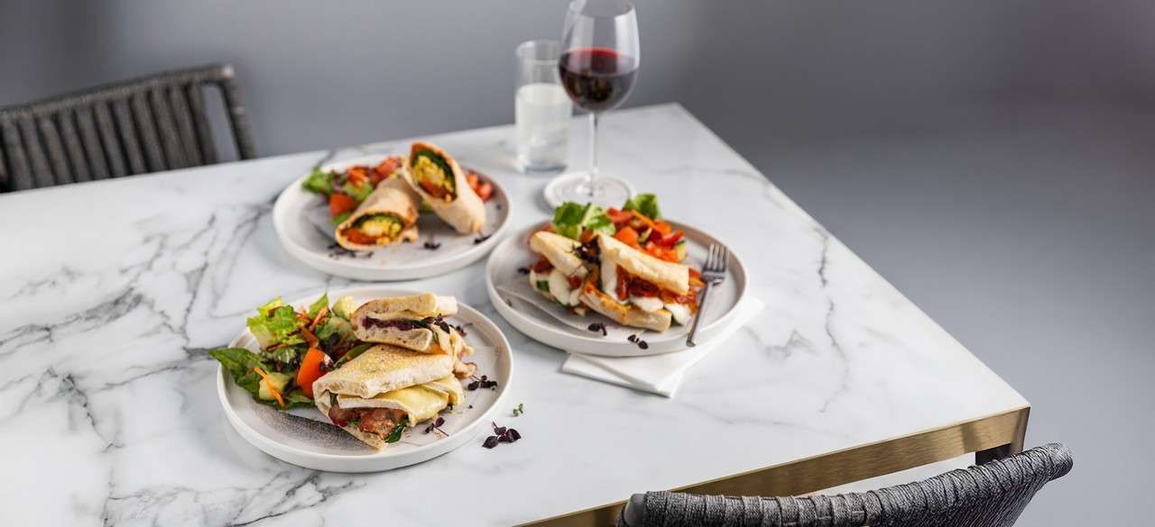 Plated sandwiches sit sliced open, revealing fillings, with mixed salads. On a marble table with utensils, a glass of red wine and water, in a minimal dining setting.
