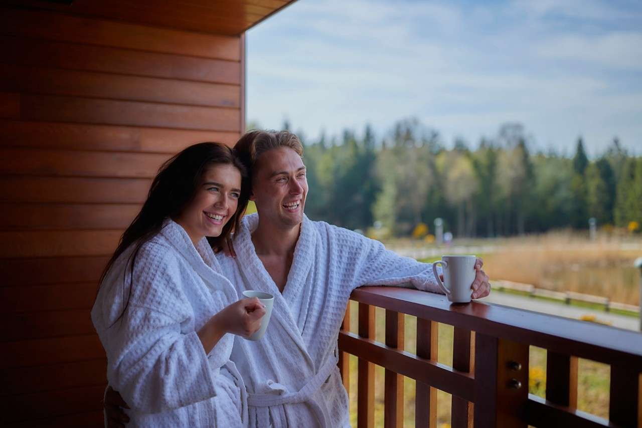 Two people in white bathrobes smile and hold mugs, leaning on a wooden balcony railing, overlooking a lakeside meadow and distant trees under a bright, partly cloudy sky.