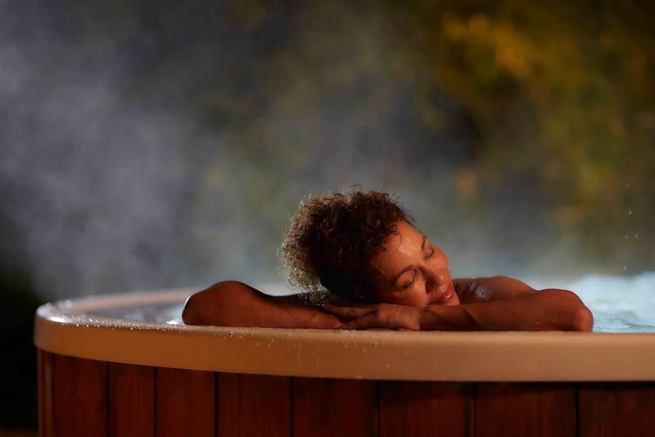 Person rests with eyes closed, leaning on the rim of a steaming wooden hot tub, tranquil. Soft night lighting and blurred foliage surround, water droplets glistening on the edge.