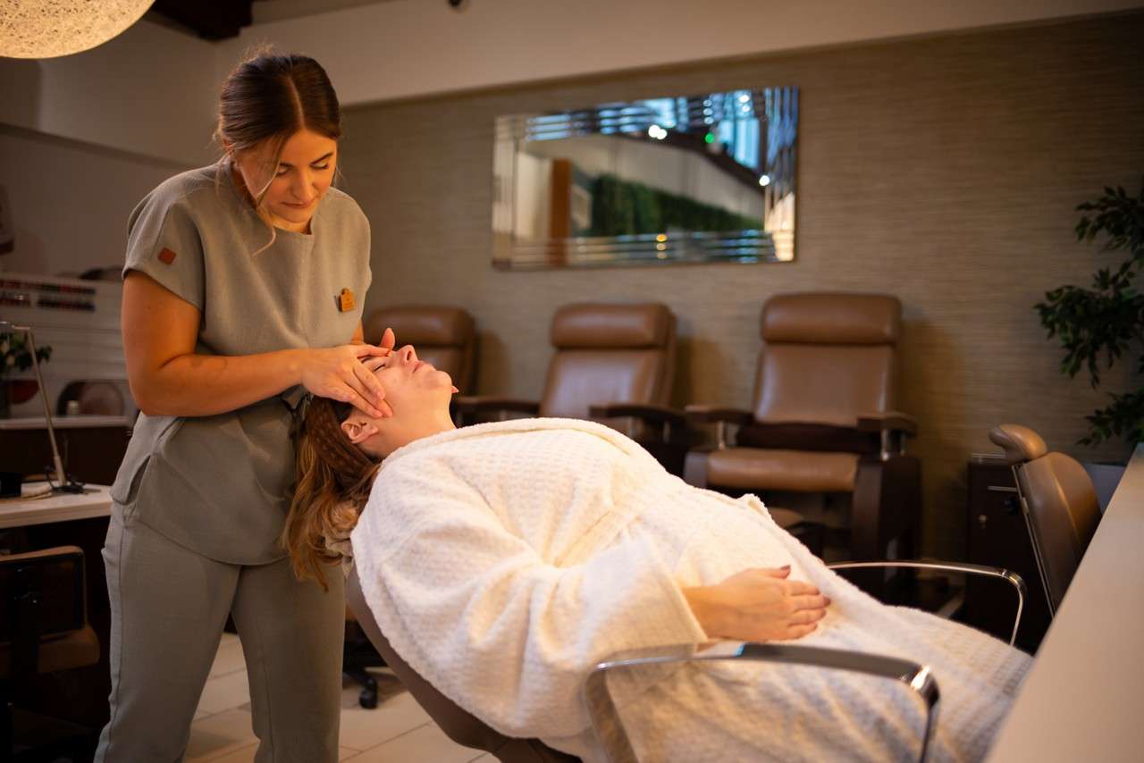 A spa therapist massages a client's face while the client reclines in a chair, wearing a white robe; warm-lit salon with leather chairs, mirror, and plants in the background.