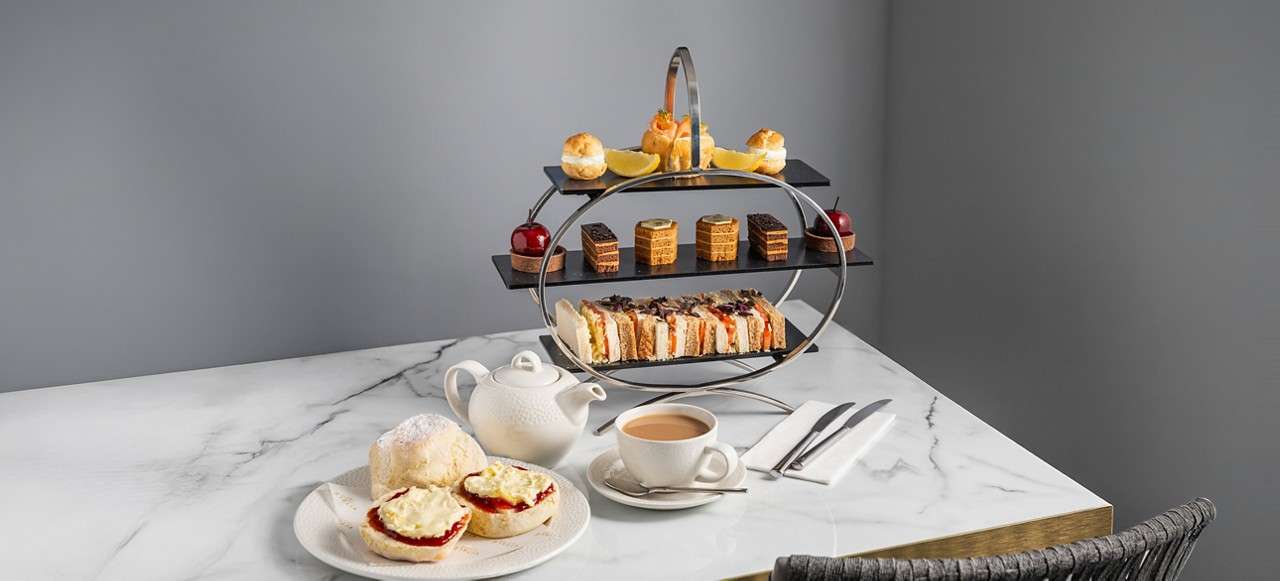 Three-tier afternoon tea stand displays sandwiches, cakes, and pastries; scones spread with jam and cream sit beside a teapot and cup on a marble table in a gray room.