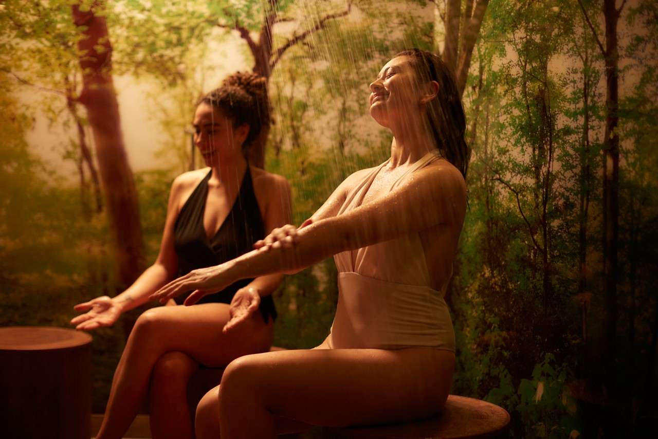Two women in swimsuits sit on round wooden stools, catching warm shower streams and rubbing arms, in a dim, spa-like room with a forest mural background and soft golden light.