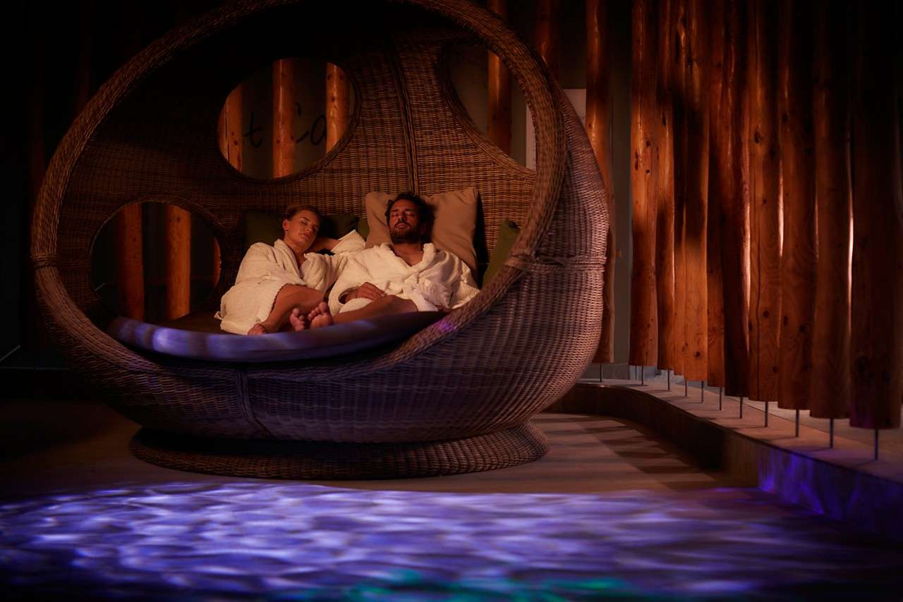 Two people recline, resting in a large round wicker pod-chair, wearing white robes, in a dim spa room with wooden walls and blue light rippling across the floor.