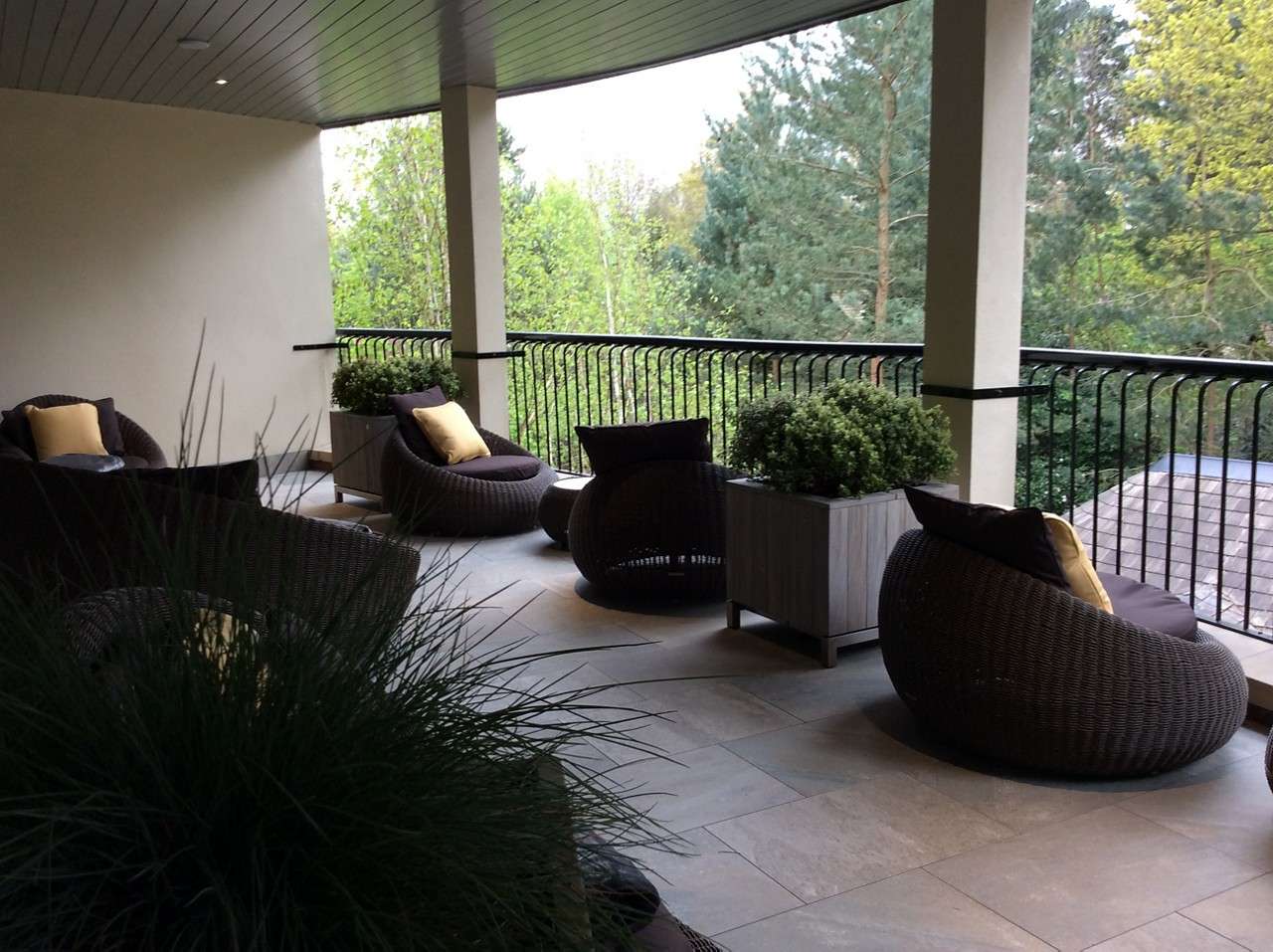 Curved wicker lounge chairs with cushions face outward, grouped around small tables, on a covered balcony. Potted shrubs line the railings, overlooking leafy trees and a rooftop.