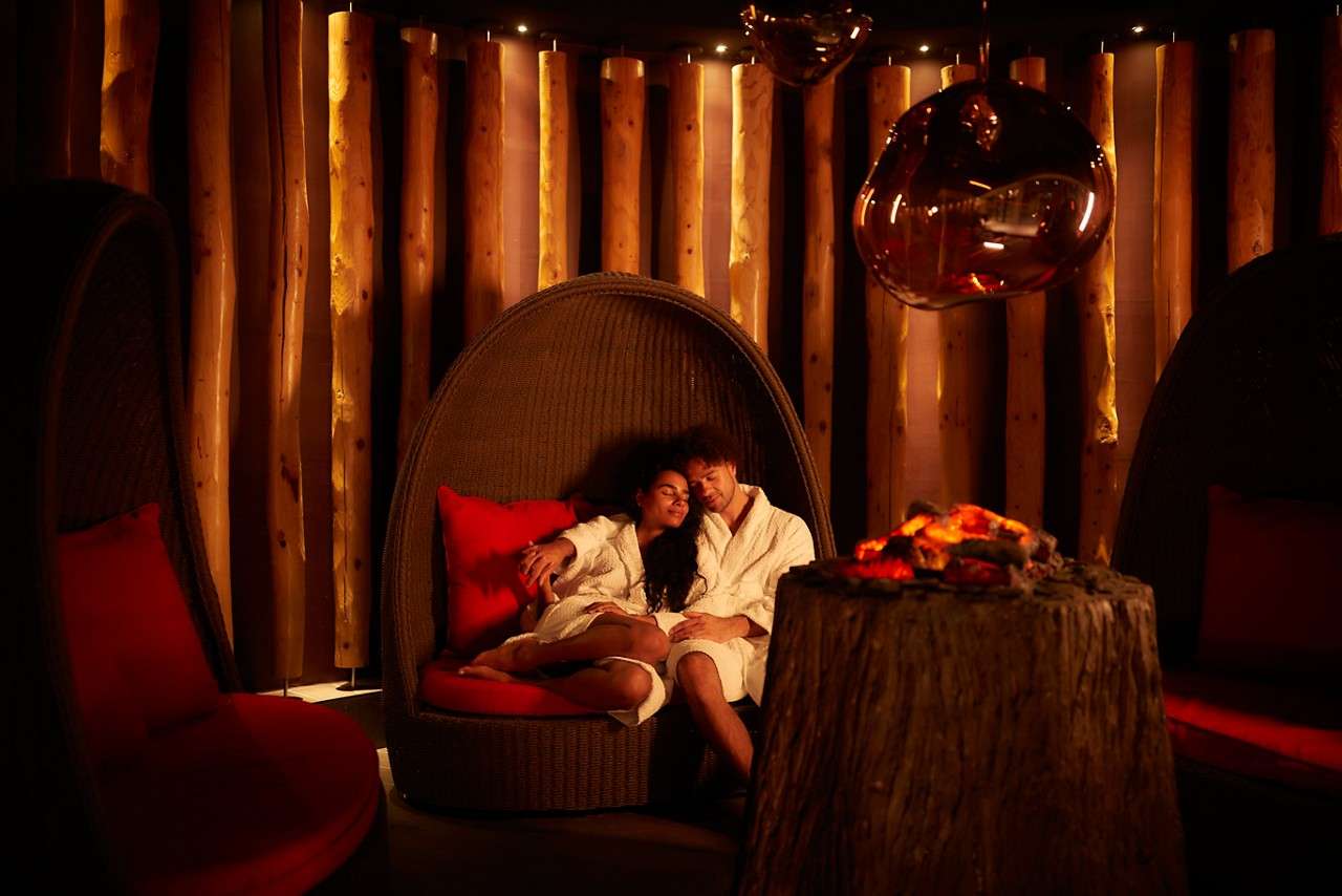 Couple in white bathrobes reclines together in a wicker pod chair, relaxing beside a glowing faux fire on a stump, amid red cushions, amber glass lights, and a log-wall backdrop.