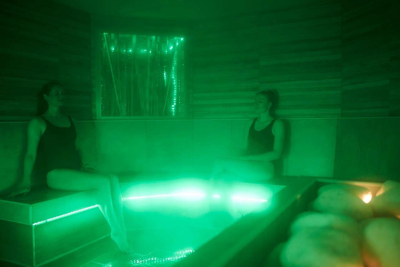 Two people in swimsuits sit relaxed on tiled benches, illuminated by green LED lights and mist, inside a spa steam room; sauna stones glow in the foreground.