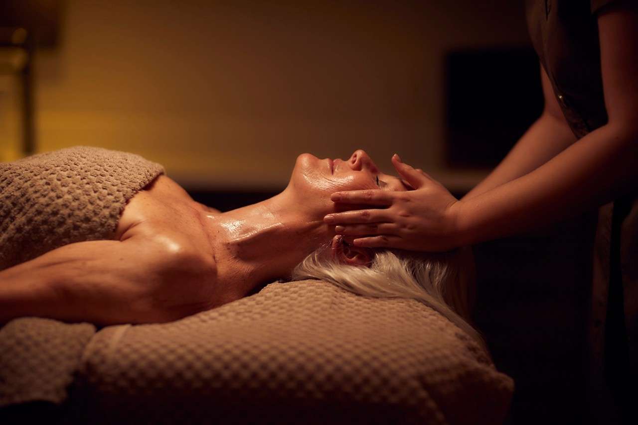 Client receives a facial massage, eyes closed, with oiled skin, while lying under a textured towel on a spa table in warm, dim lighting.