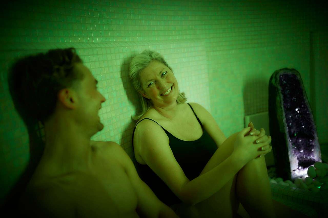 Two people in swimsuits relax and chat, seated against tiled walls under green light, beside a glowing amethyst geode and white stones in a spa or steam room.