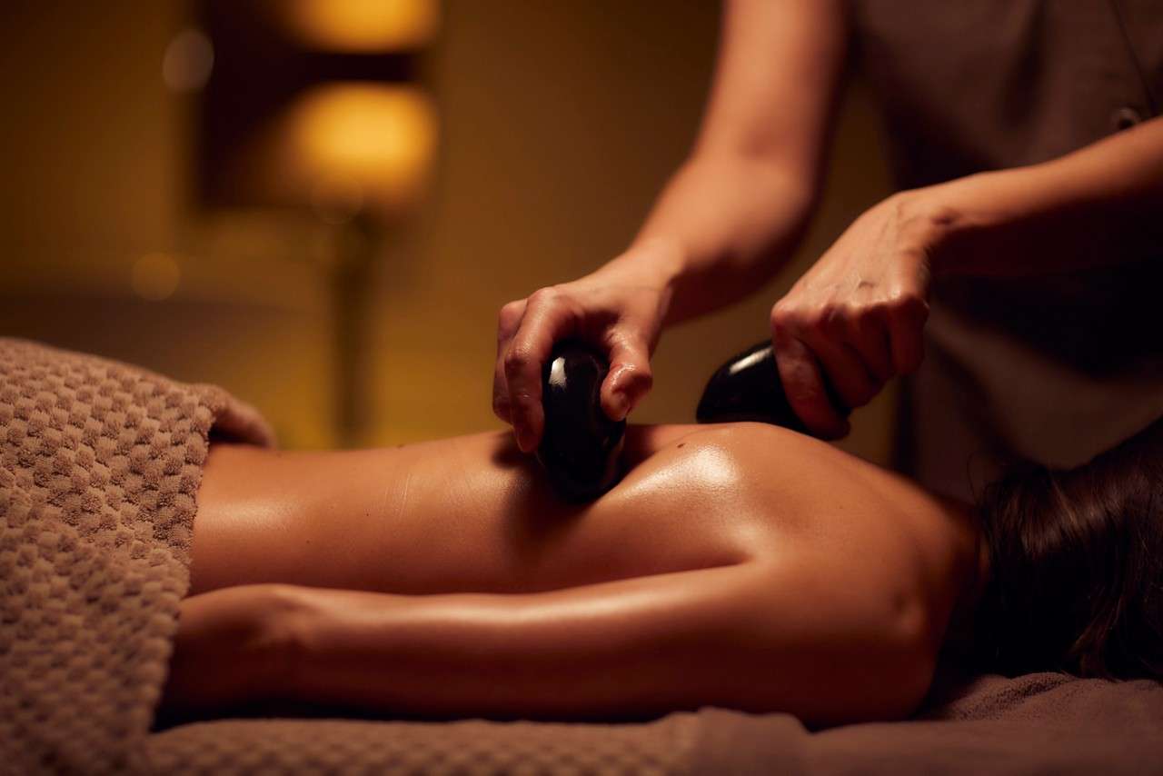 Massage therapist’s hands press hot stones across a person’s oiled back, kneading muscles; warm, dimly lit spa room with soft towels and a blurred lamp in the background.