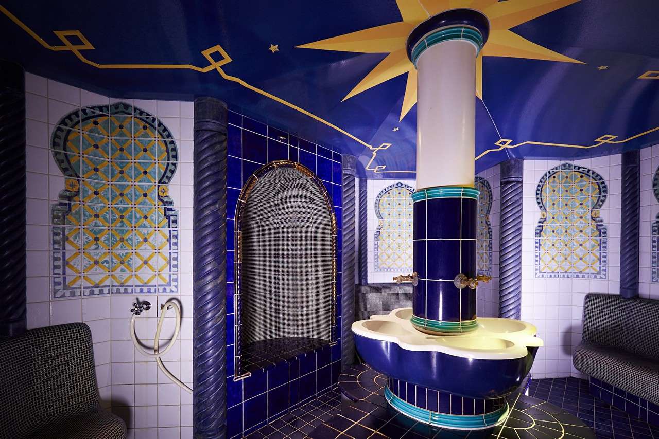 Central tiled fountain column stands with faucets feeding a scalloped basin, surrounded by blue mosaic walls, arched niches, cushioned benches, and a star-decorated ceiling in a Moroccan-style steam room.