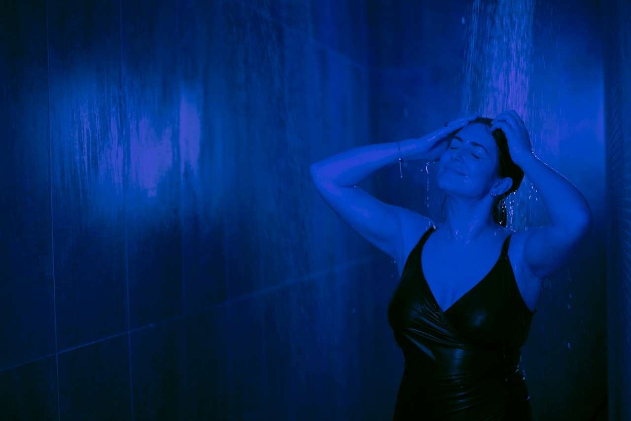Person washes hair under a running shower, eyes closed and hands massaging scalp, while water cascades down in a dark tiled stall lit by deep blue light.