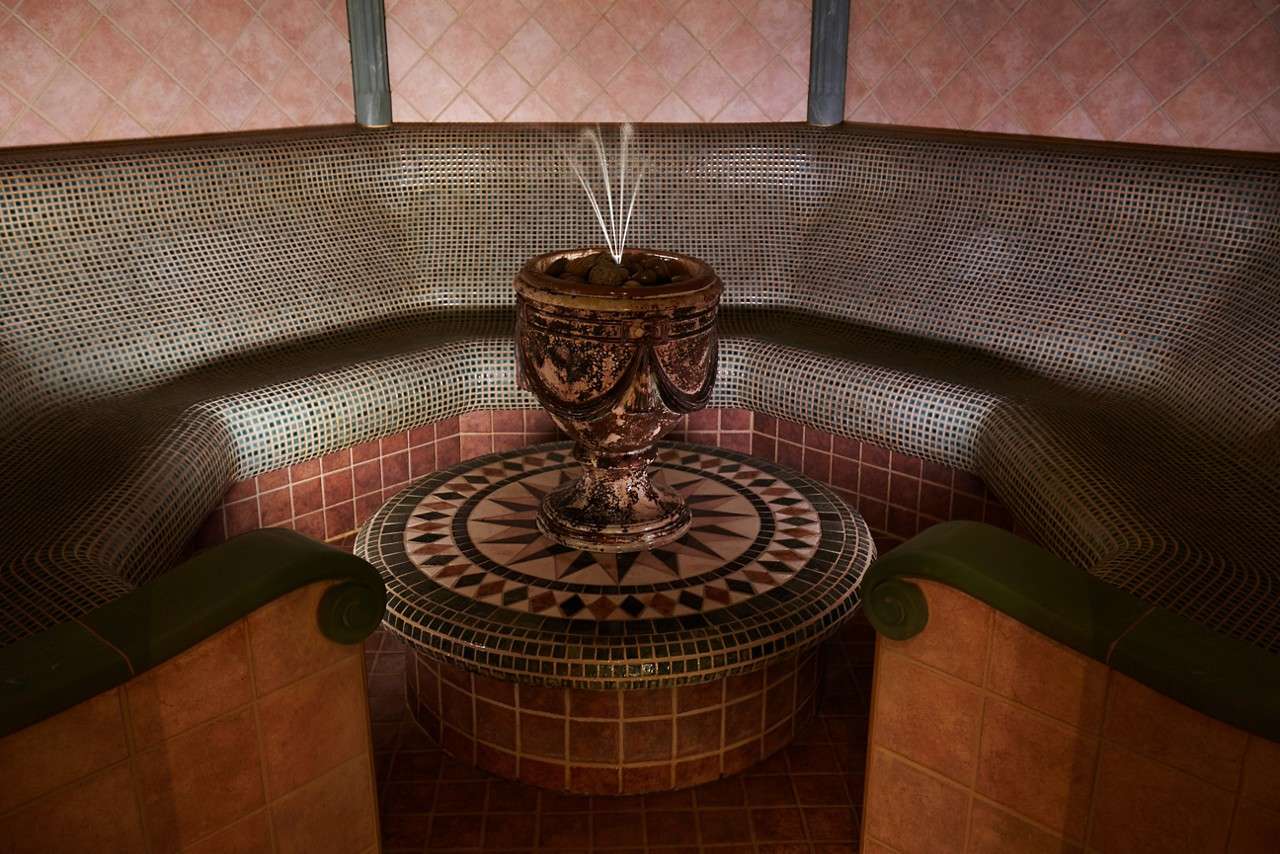Ornate urn fountain sprays thin water jets upward, resting on a mosaic pedestal. Surrounding it, a curved, tile-covered bench forms a circular steam-room alcove with warm, dim lighting.