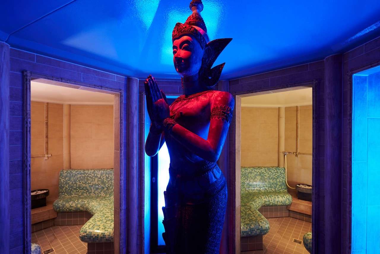 Statue with hands pressed together stands centered, illuminated blue-red, between two open tiled steam rooms. Mosaic benches curve along walls; small stone heaters sit in corners under warm beige lighting.