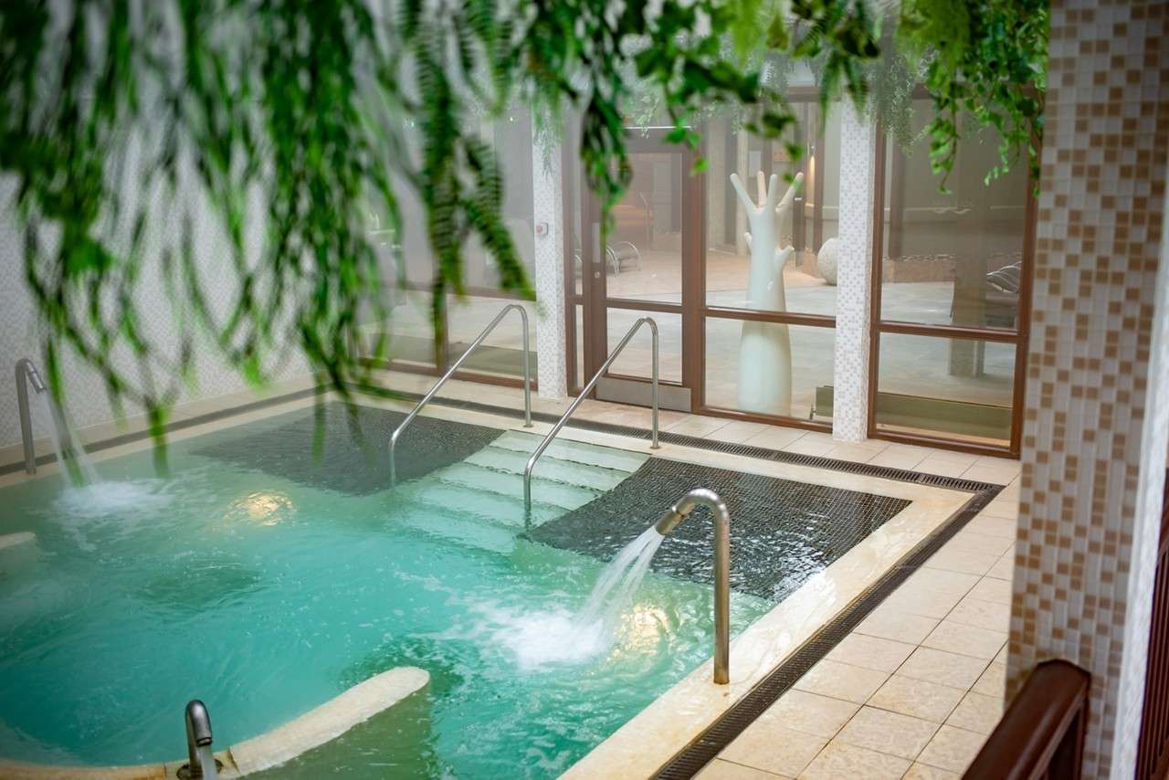 Thermal spa pool bubbles and pours from stainless spouts, with steps and handrails leading in; surrounding tile deck, glass wall, and hanging greenery reveal lounge area and white tree-shaped sculpture.