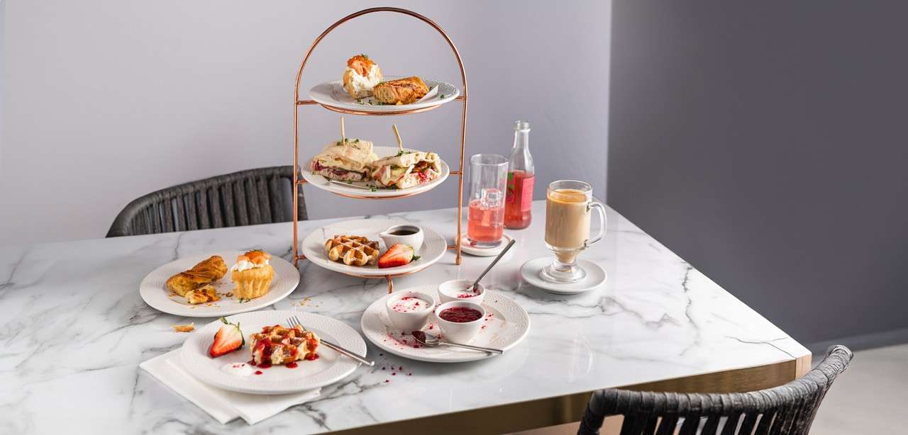 Tiered afternoon-tea stand displays sandwiches and pastries, accompanied by waffles, jam, sliced strawberries, a latte, and pink soda. Set on a marble table in a minimalist café with wicker chairs.