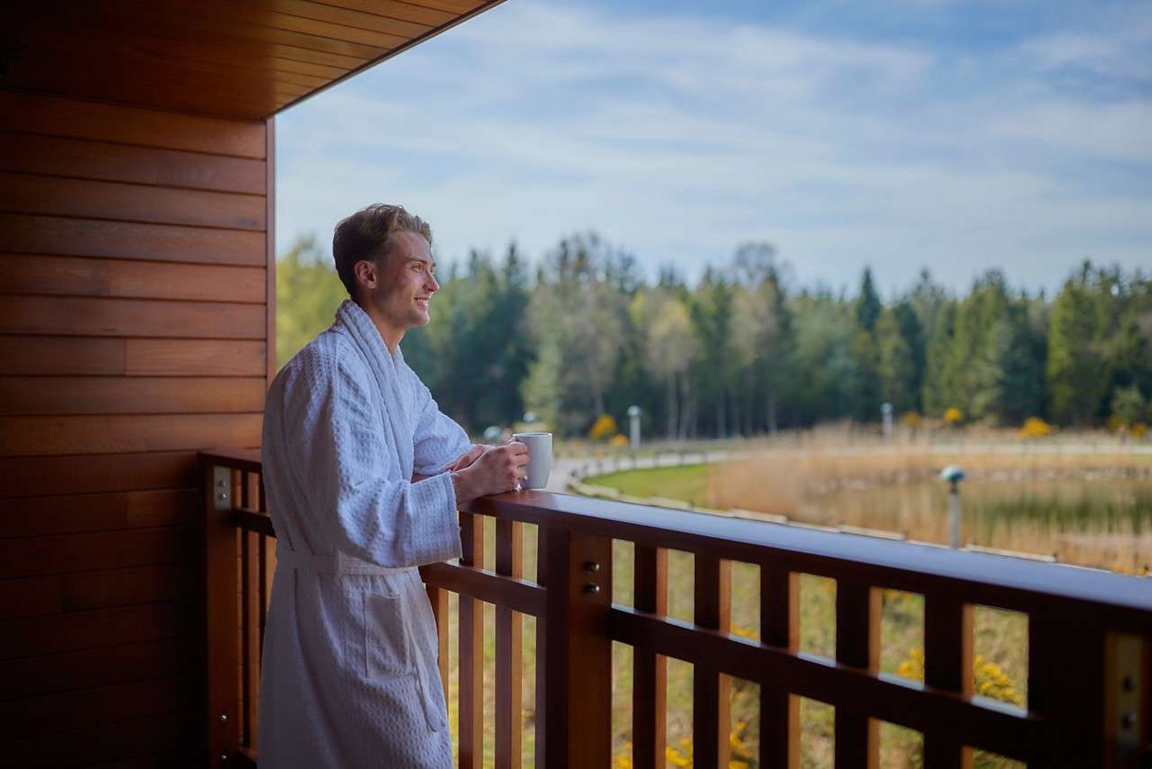 Man in a white bathrobe holds a mug, leaning on a wooden balcony railing, overlooking a peaceful landscape with trees, a pond, and a curving path under a blue sky.