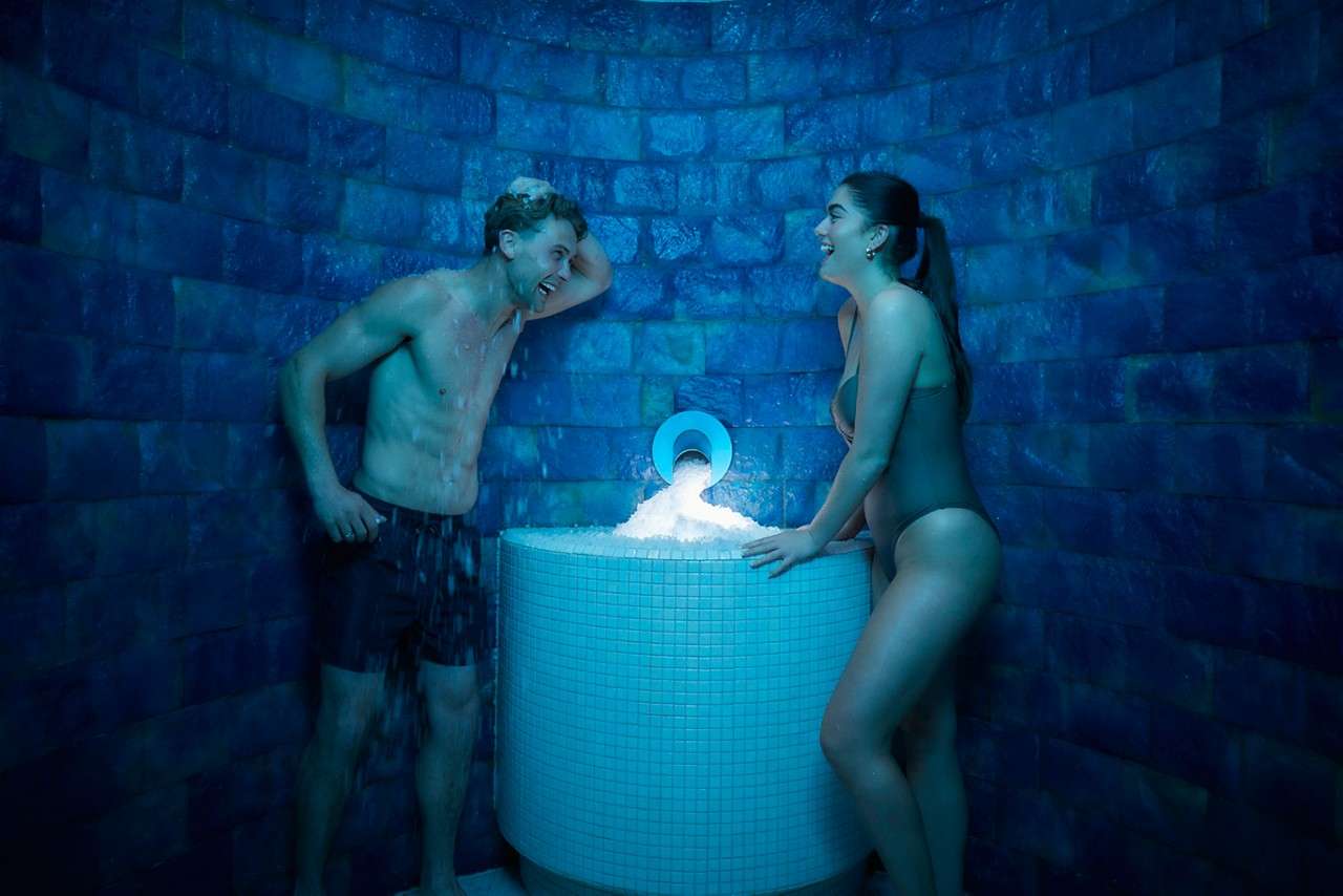Two swimsuit-clad adults gather ice from a glowing dispenser, laughing under water spray, inside a blue, circular tiled spa chamber.