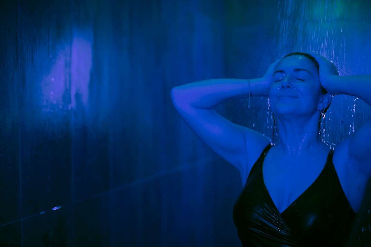 Person in a black swimsuit stands under a shower, eyes closed, hands on head, enjoying water. Set within a dark, tiled shower room illuminated by blue-green light.