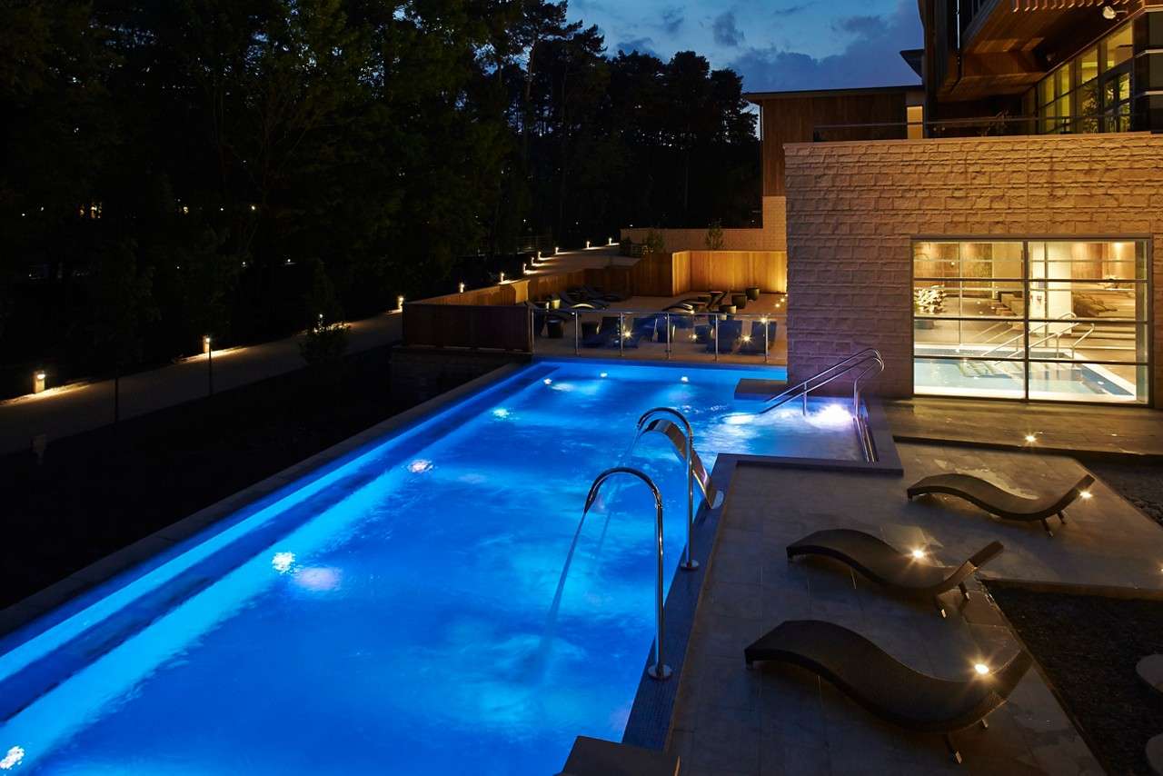 Swimming pool glows blue, jets arching water; nearby sun loungers sit empty, while a modern spa building and glass windows overlook pathway lights and surrounding trees at dusk.