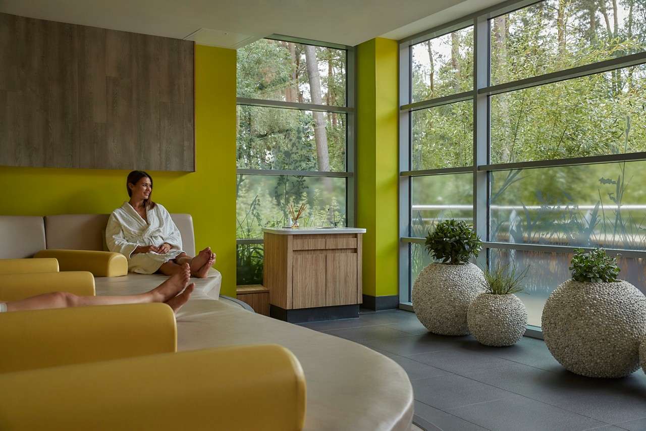 A person in a white robe relaxes on a padded lounge chair, in a bright spa room with lime-green walls, large windows, spherical planters, and forest greenery visible outside.