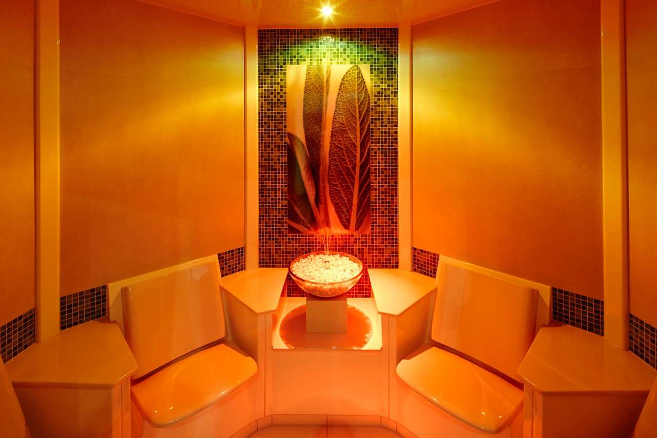 Bowl of white stones rests on a pedestal, centered between two molded benches, as warm orange lights bathe a tiled alcove with large leaf artwork, creating a symmetrical spa-like room.
