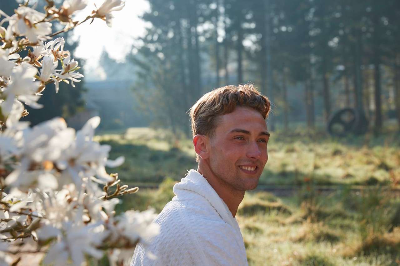 A young adult smiles while turning toward the camera, wearing a white robe, in morning light amid white blossoms, with a grassy field and tall trees in the background.