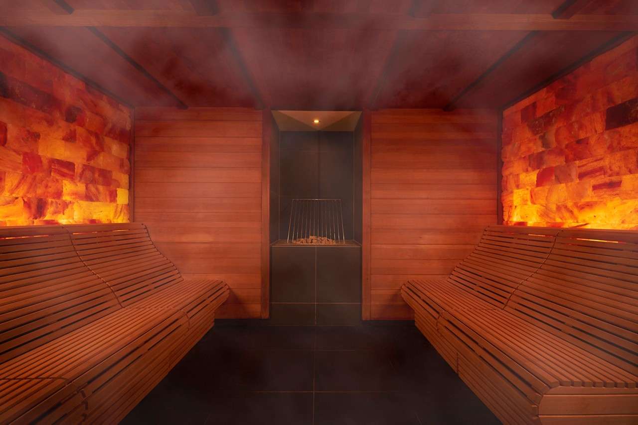 Wooden sauna benches line both sides, steaming gently; a central heater holds hot stones. Warm backlit salt-brick walls and wood paneling create an enclosed, dim, serene room.