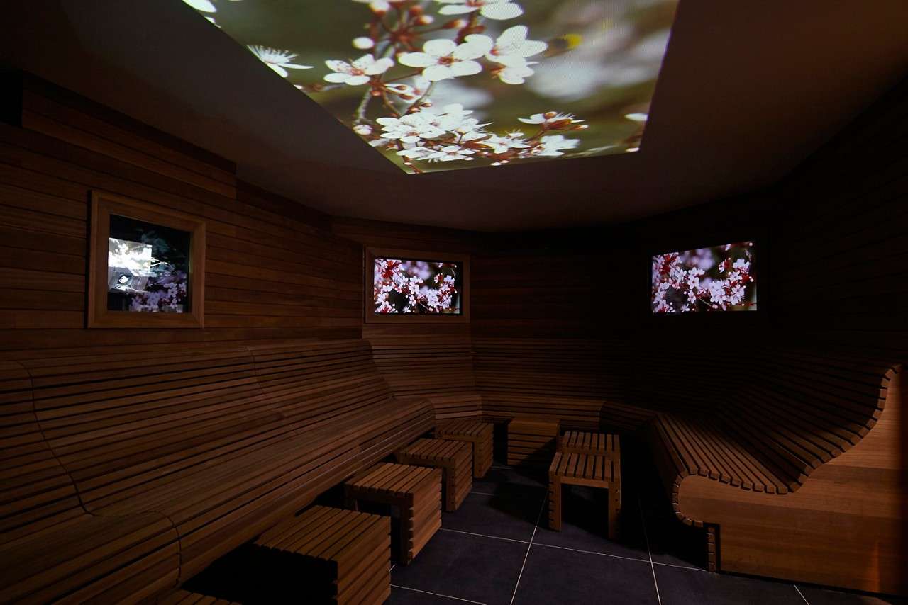 Ceiling and wall screens display cherry-blossom visuals as wooden bench seating lines a dim spa room, with slatted stools on dark tiled flooring creating a tranquil, enclosed lounge.