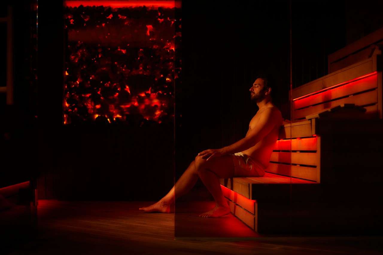 Man sits relaxing with eyes closed, leaning against wooden tiered bench, legs extended. Warm red lighting glows beneath benches and wall panel, suggesting a dim sauna or infrared heat room.
