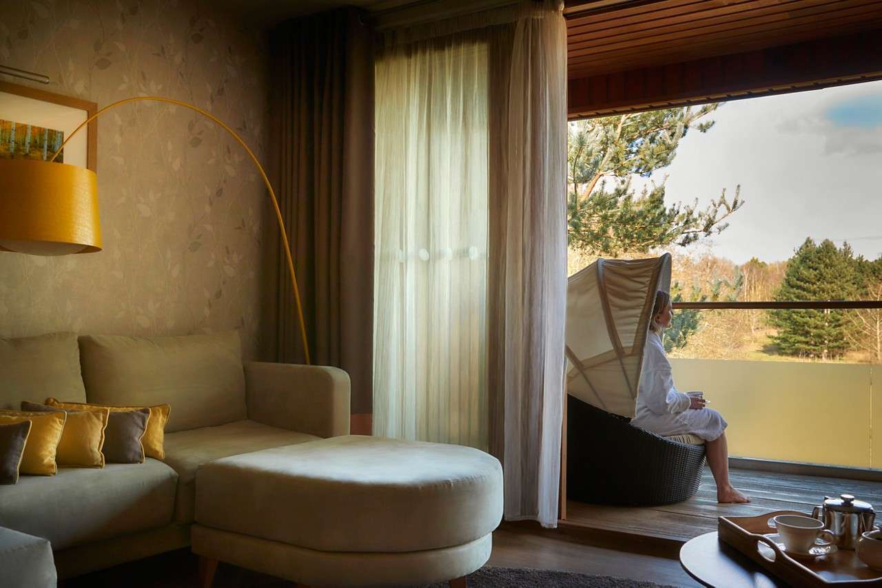 A person in a robe sits on a patio chair, sipping from a cup, on a balcony overlooking trees, beside a living room with sectional sofa, lamp, and sheer curtains.