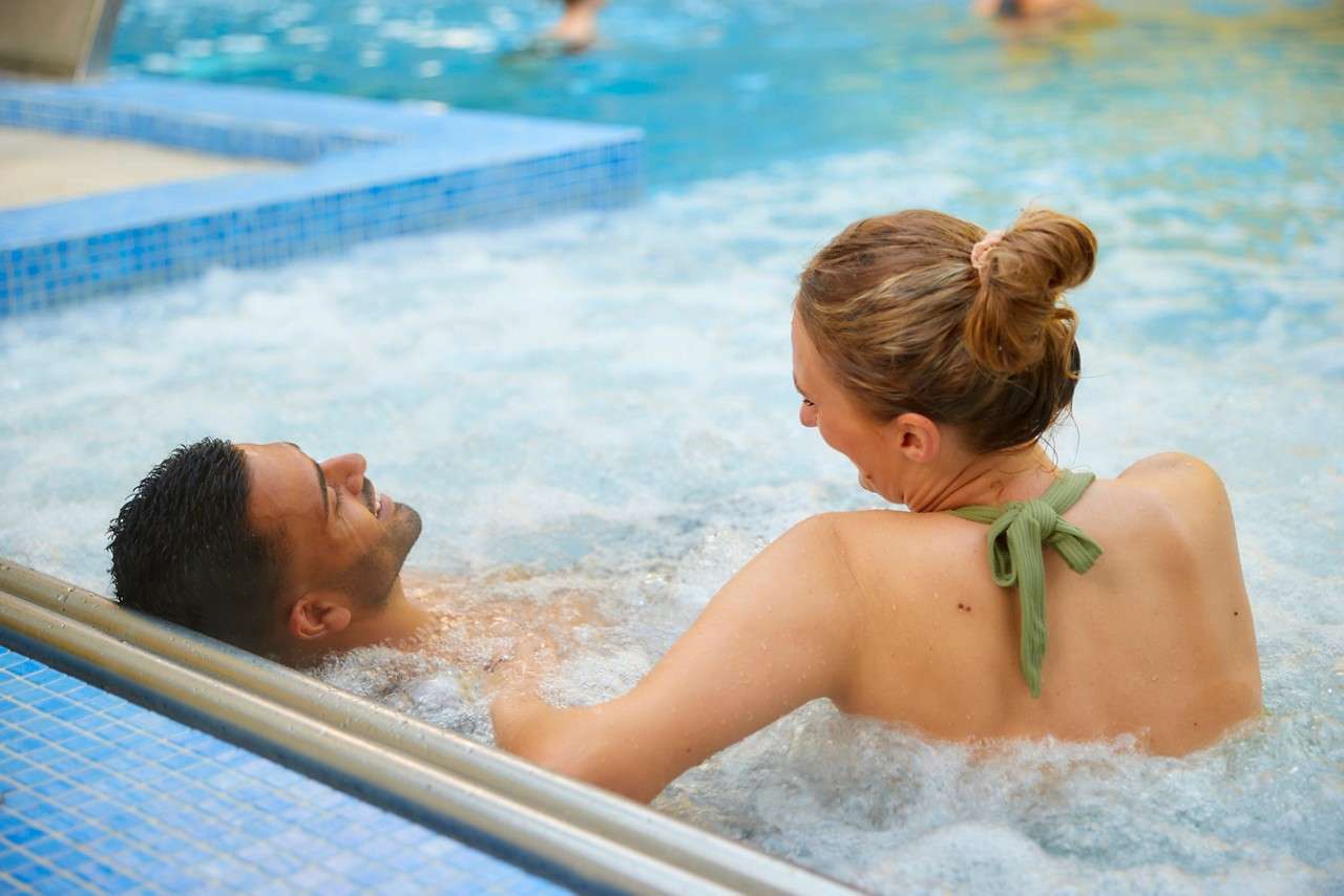 Two adults relax and chat while sitting in bubbling water, one reclining against a metal edge in a tiled spa or pool area with blue water and distant swimmers.