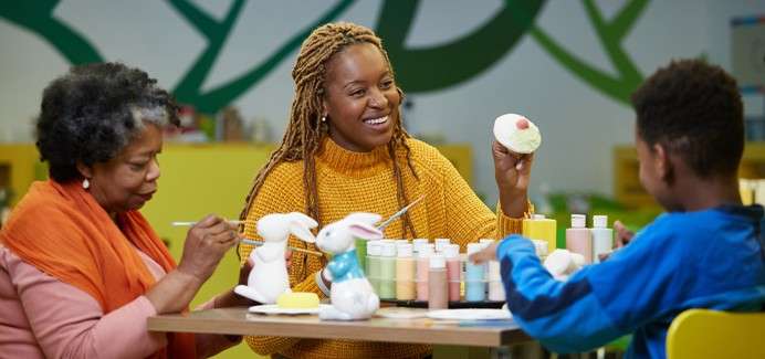 Three people paint ceramic rabbit figurines, using brushes and pastel paints, around a table. A woman holds a painted donut-shaped piece. Context: craft studio with green wall patterns and supplies.