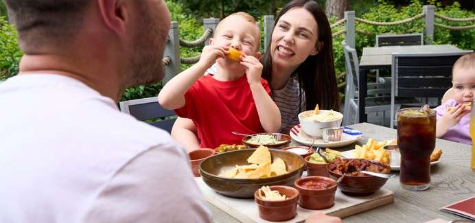 Child eats nachos while seated with parents at an outdoor restaurant table; bowls of dips, fries, soda, and cutlery on a wooden table; greenery and empty chairs in the background.