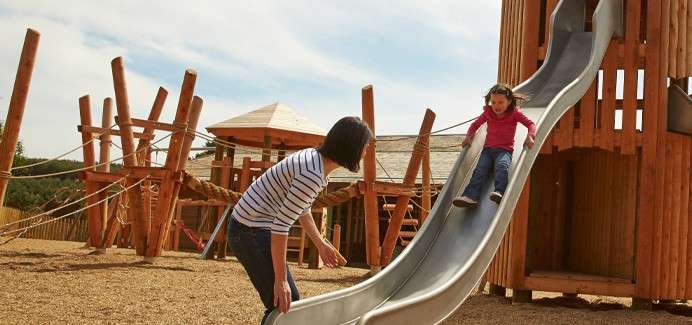 A woman stood at the bottom of a slide whilst a little girl slides down.