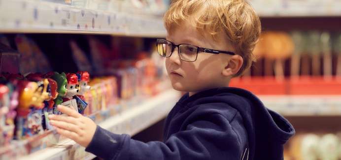 Child with glasses reaches to examine small toy figures on a store shelf, standing in a retail aisle with other colorful items blurred in the background.
