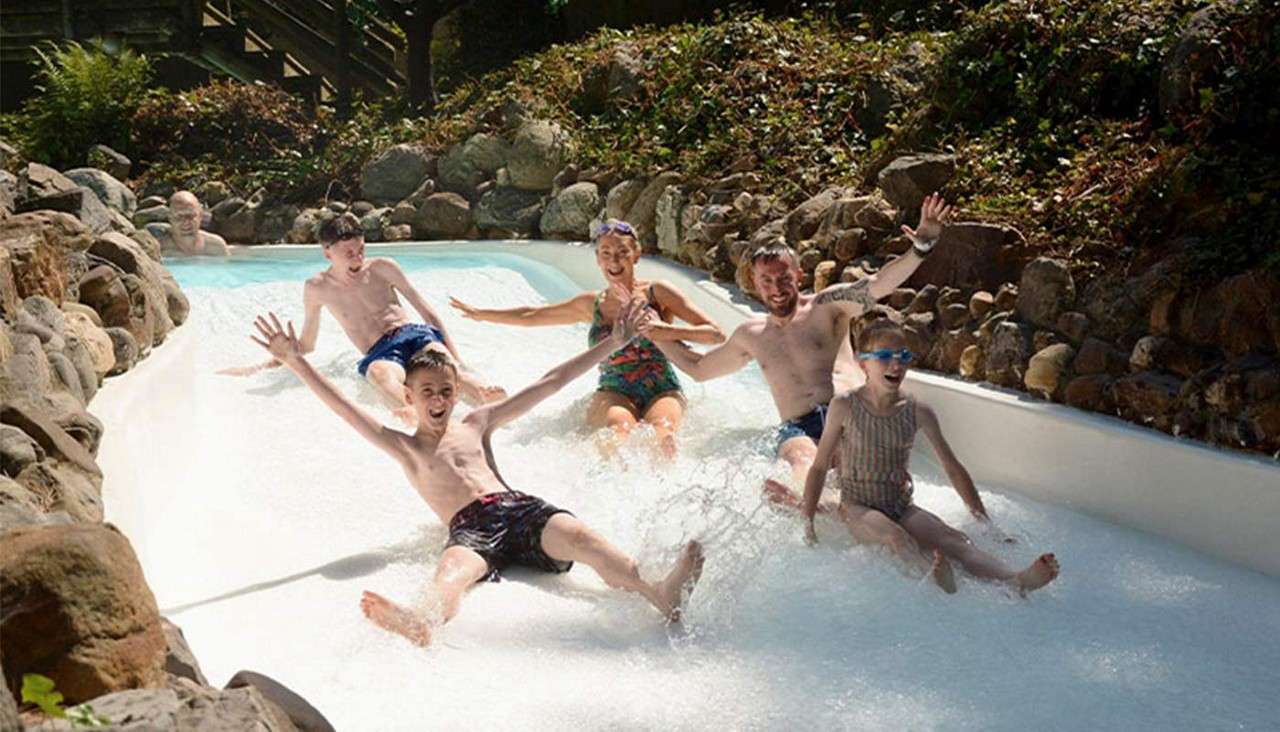 Group of swimsuit-clad people slide feet-first and splash down a shallow water chute, arms raised, in an outdoor water park channel bordered by rocks and greenery.
