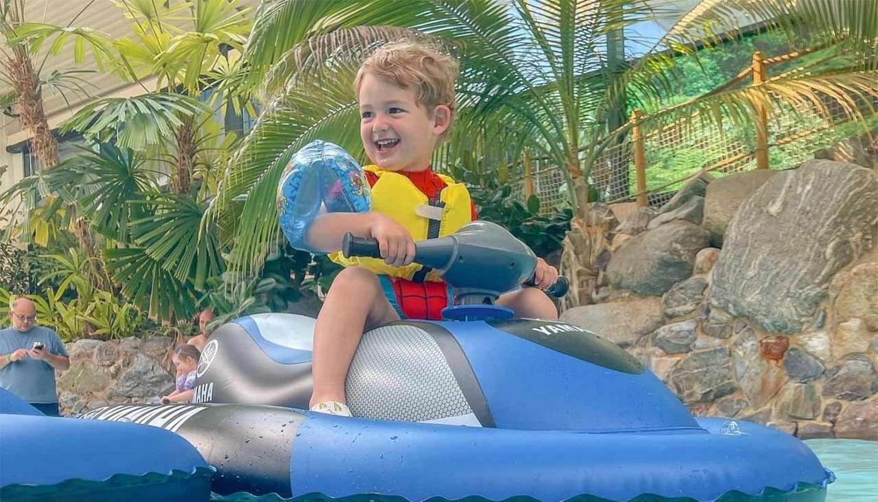 Child rides an inflatable jet ski, smiling and gripping handlebars, wearing a yellow life vest and blue arm floaties; in a pool surrounded by tropical plants, rocks, and onlookers. Text: YAMAHA, YAMAHA.