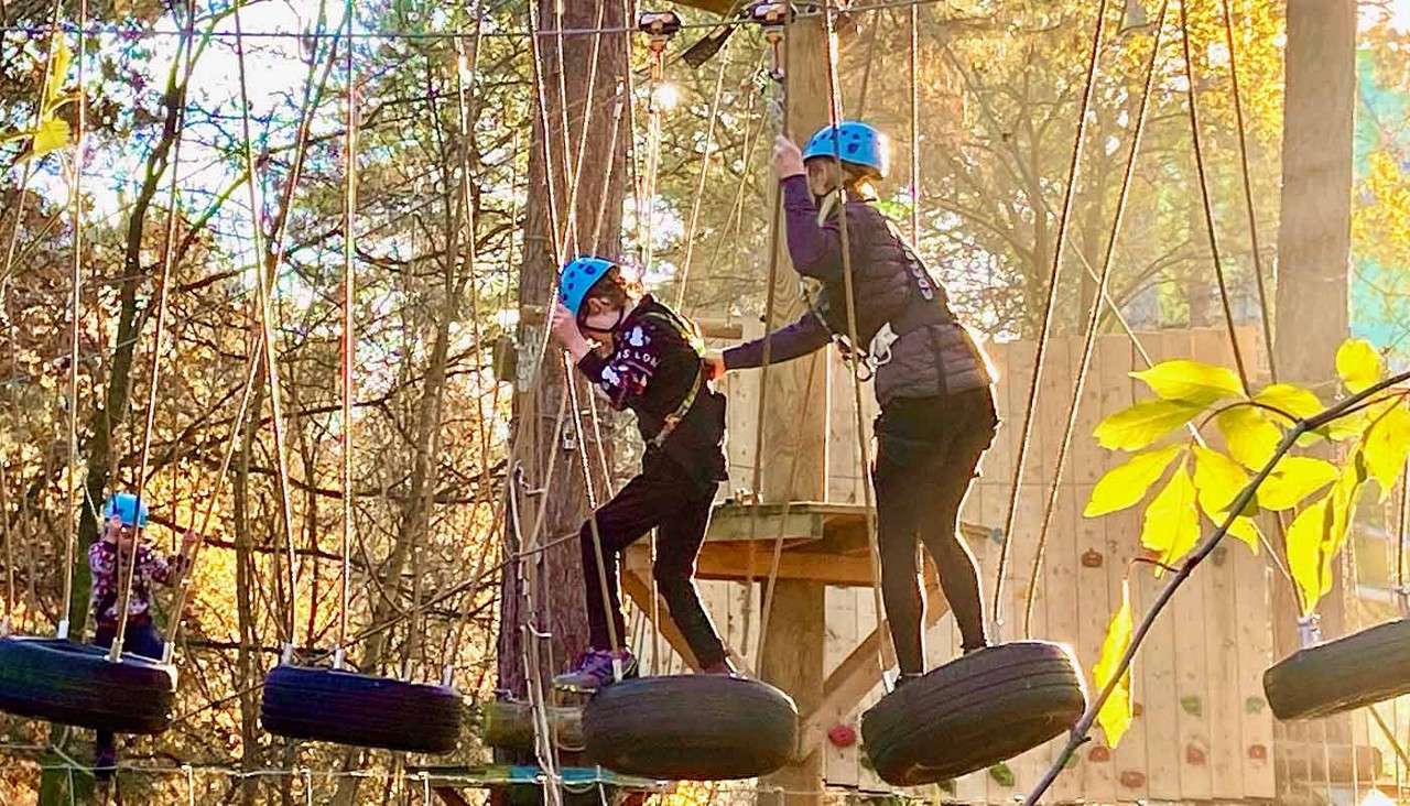 Helmeted climbers step across swinging suspended tires, clipped to overhead safety lines, as they navigate a treetop ropes course among tall trees and wooden platforms in warm autumn light.