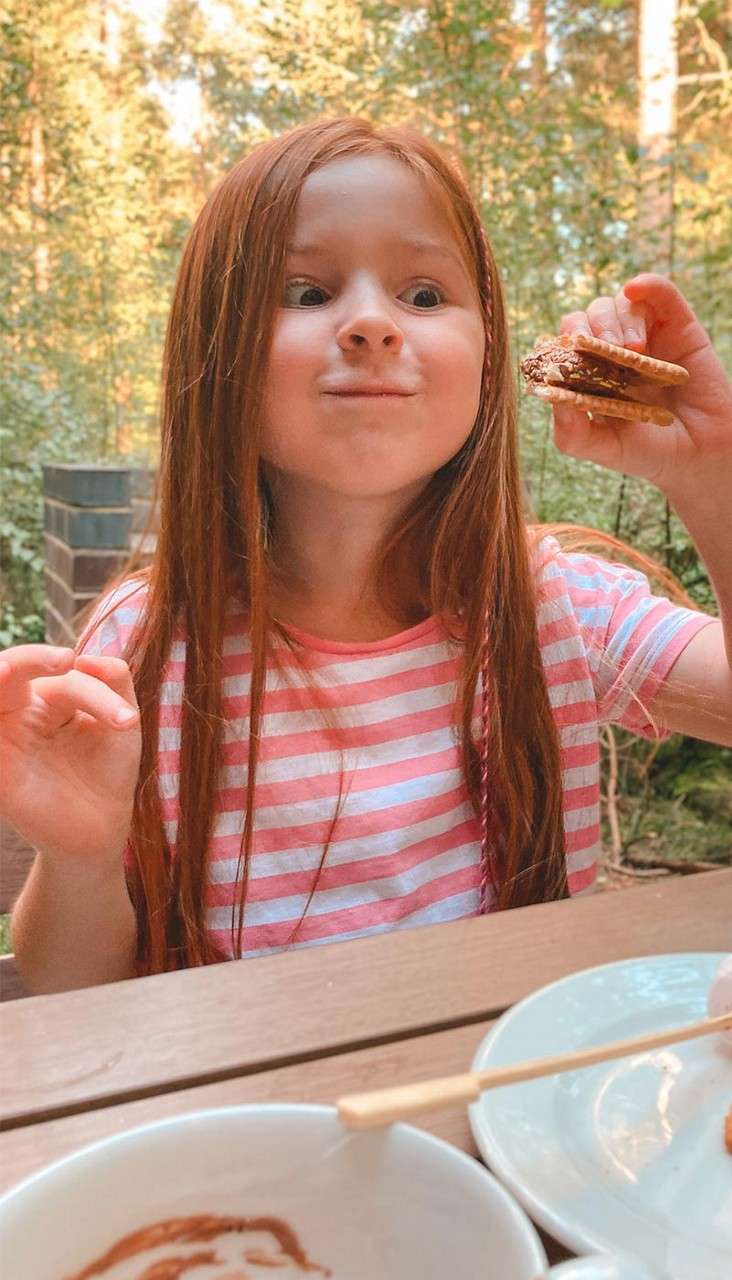 Child with long red hair holds a cookie-chocolate sandwich, puffing cheeks playfully, at a wooden picnic table with dishes in a sunlit forest setting.
