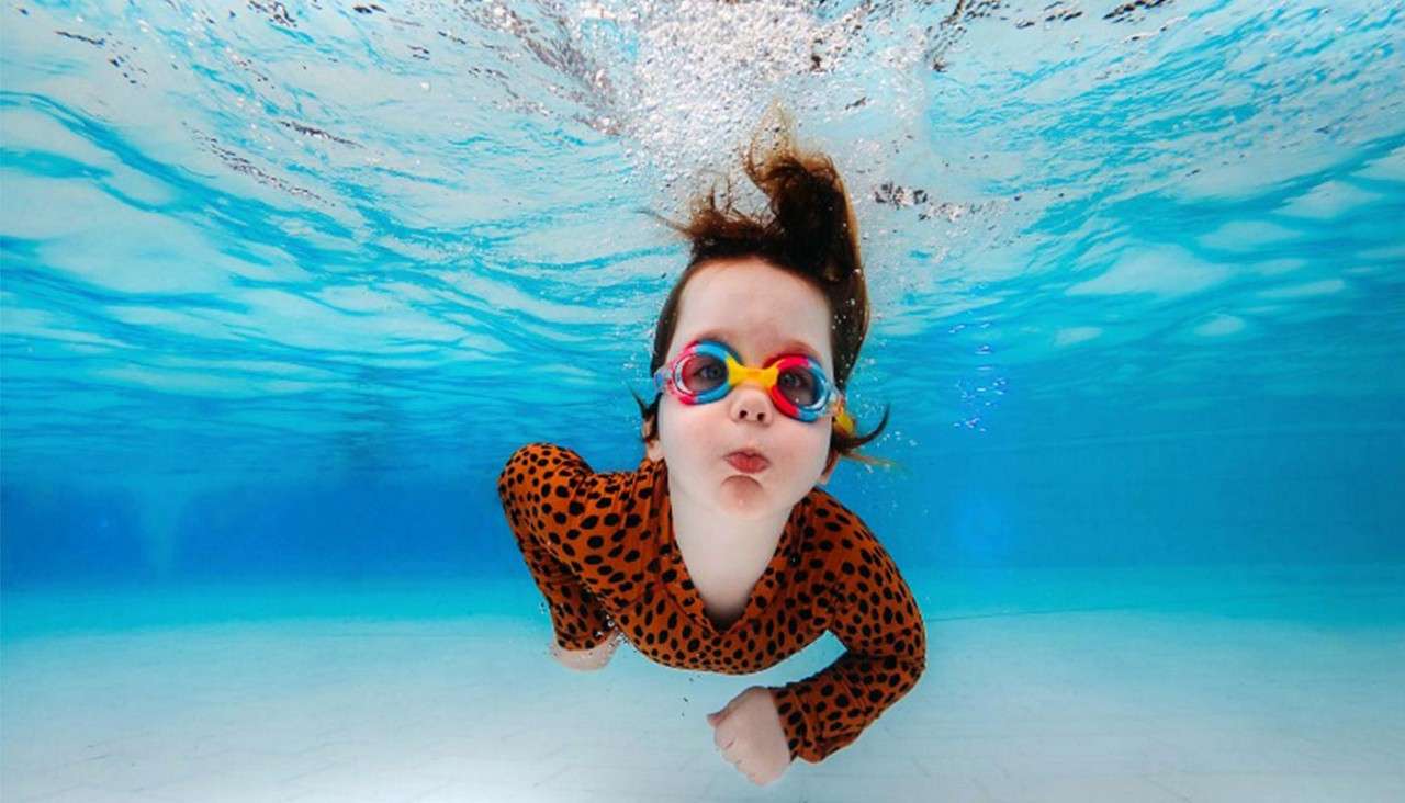 Child in multicolored goggles swims underwater, cheeks puffed, arms moving forward, in a bright blue swimming pool, wearing an orange long-sleeve black-spotted swimsuit; bubbles trail above.