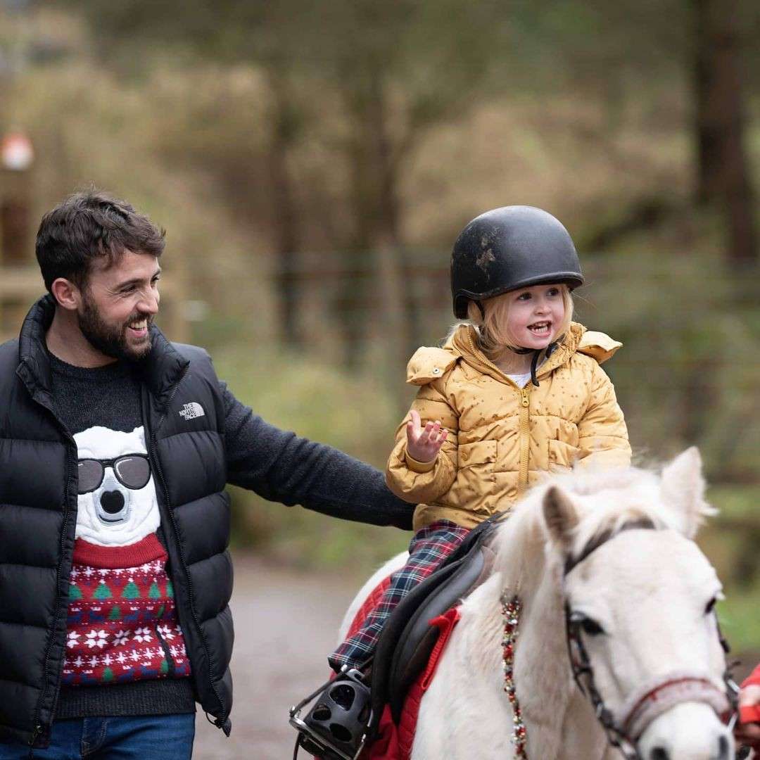 Child on a small white pony rides forward, smiling and gesturing, while an adult walks beside guiding. Outdoors on a wooded path. Visible text: THE NORTH FACE on the adult’s jacket.