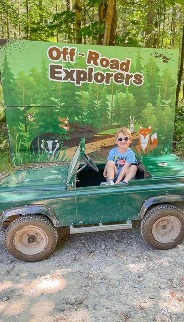 Child sits in a small green off-road toy jeep, smiling with sunglasses, parked on gravel before a forest-themed sign reading: Off - Road Explorers, in a wooded outdoor setting.