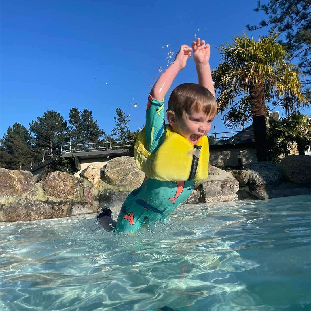 Child wearing a yellow life vest leaps and splashes with arms raised, mouth open; in a shallow pool surrounded by rocks, palm trees, and blue sky.