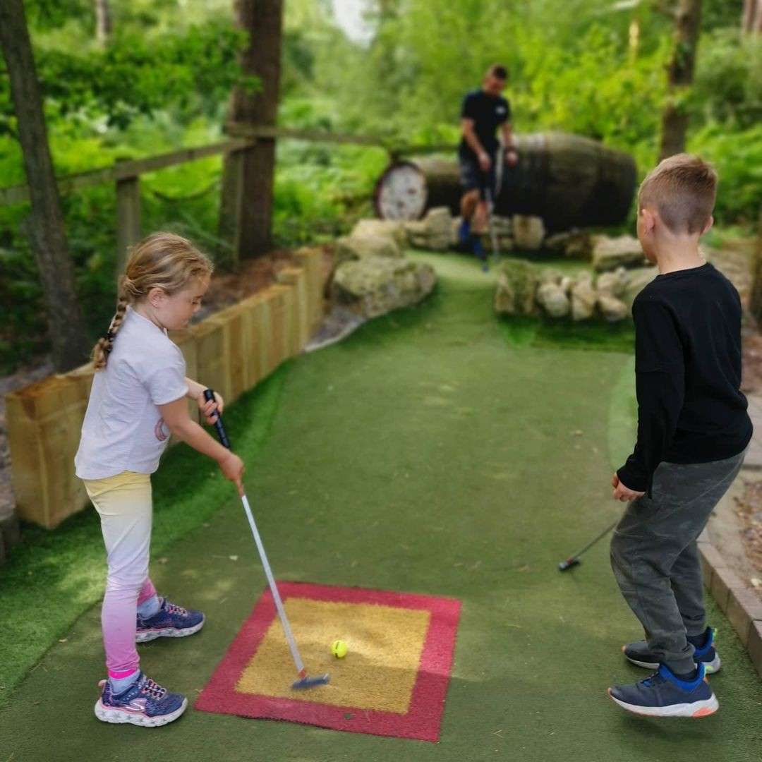 Girl golfer putts a yellow ball from a marked mat, while a boy watches. In the background, two players handle clubs near a barrel obstacle on a wooded mini-golf course.