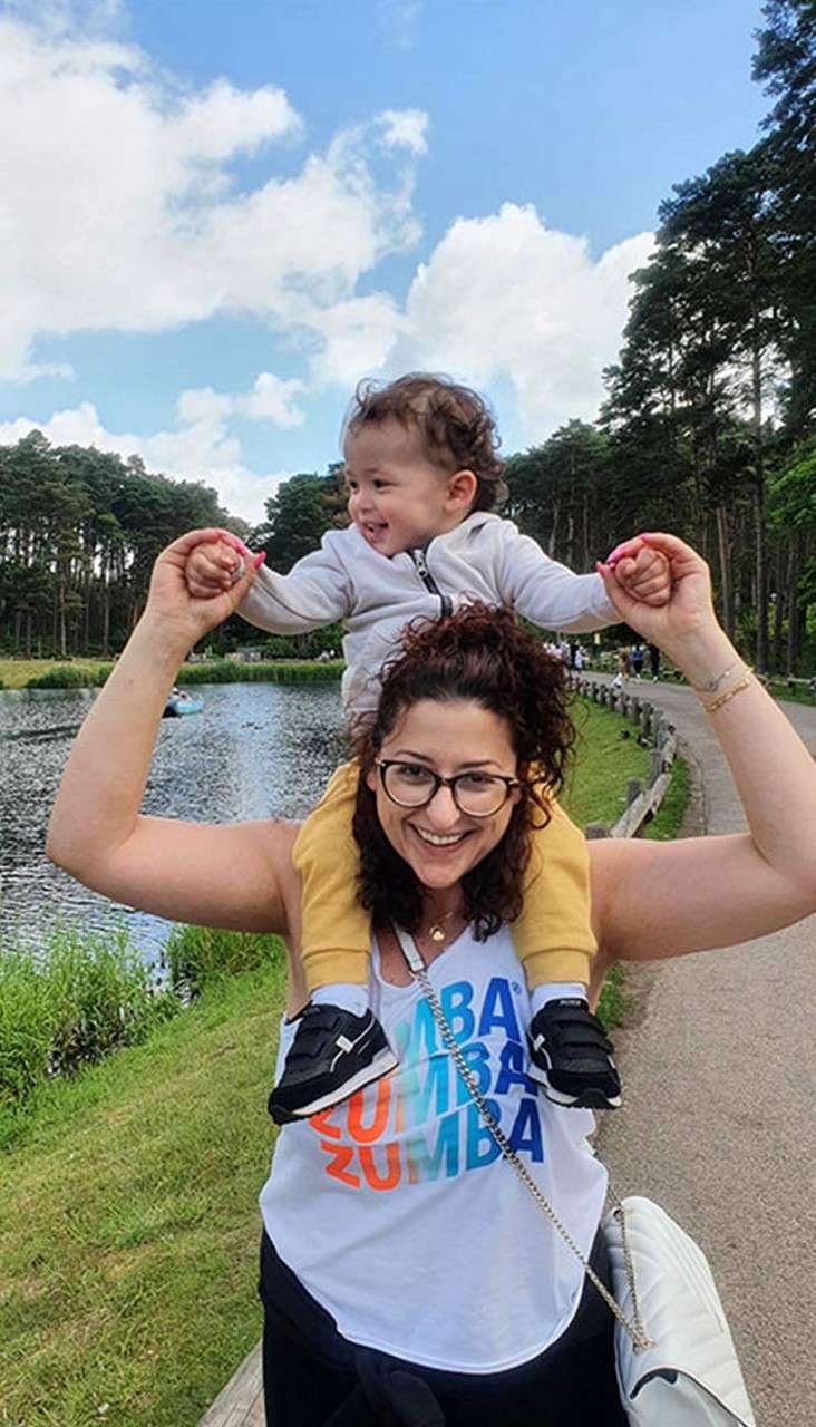 Toddler sits on a woman’s shoulders, raising arms; lakeside path with trees and blue sky. Shirt text: ZUMBA ZUMBA ZUMBA ®.