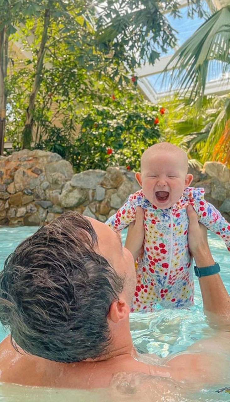Baby with eyes closed and mouth open is lifted by an adult in a pool, surrounded by rocks, palm trees, and greenery under a bright glass-roofed atrium.